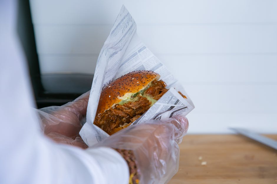 Close-up of a gloved hand holding a pulled pork burger wrapped in printed paper